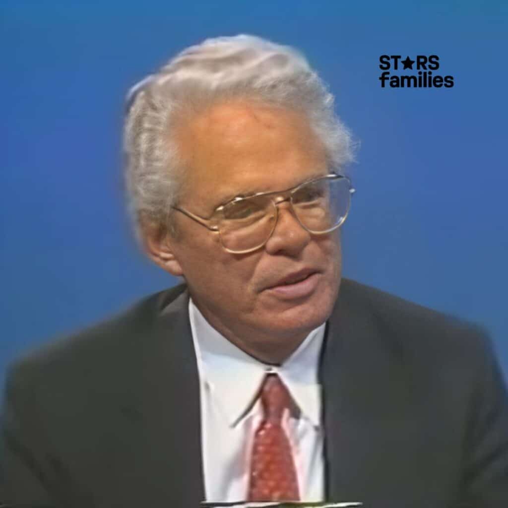 John Teets, wearing a suit and tie, is positioned against a solid blue background.