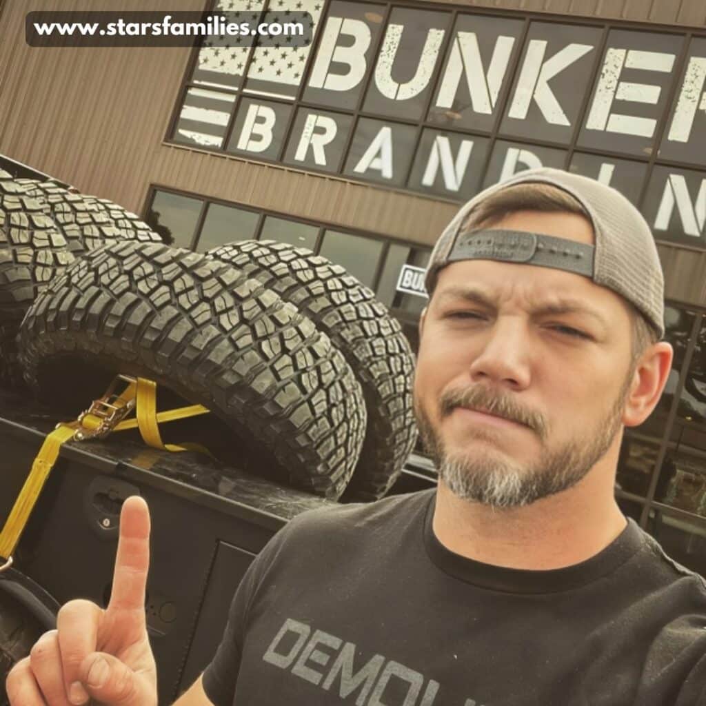 Matt Carriker, wearing a black shirt with the word "DEMOL" and a cap, stands in front of a building with a sign that reads "BUNKER BRANDING." He is pointing upwards with one finger. The background includes large off-road tires secured with yellow straps on a vehicle.