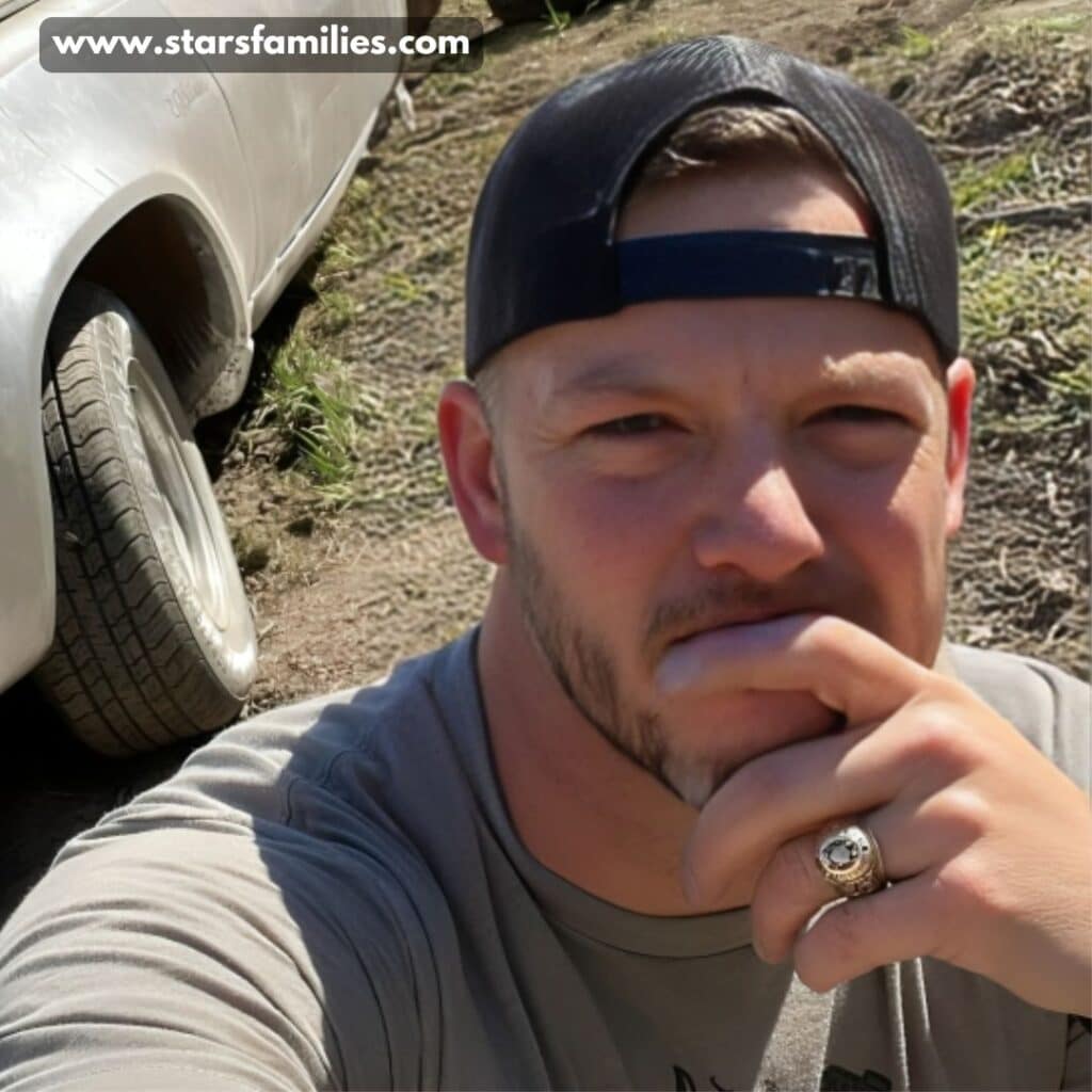 Matt Carriker, wearing a black cap and a gray shirt, is seen holding a hand with a ring near his chin. He is standing next to a white vehicle with a flat rear tire, parked on a dirt surface with some grass.