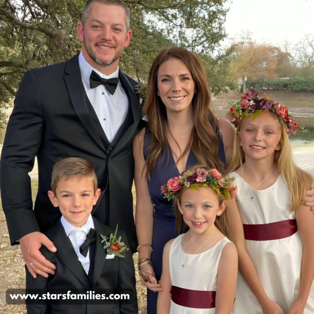 Matt Carriker, wearing a black tuxedo with a bow tie, poses outdoors with his family. His wife is in a dark blue dress, and their three children are dressed formally, one boy in a black suit and two girls in white dresses with maroon sashes and flower crowns.