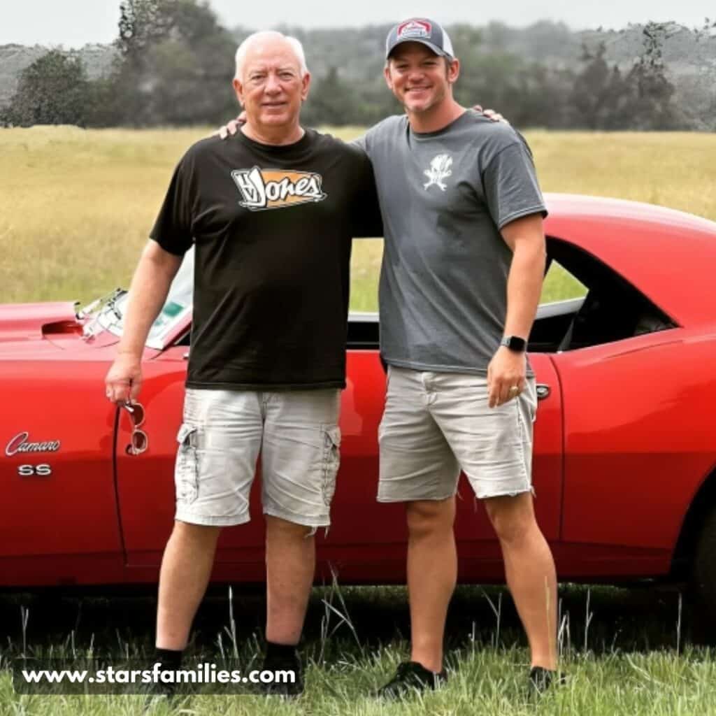 Matt Carriker, wearing a black t-shirt with "Jones" printed on it and light-colored cargo shorts, stands beside his father(Dr. Lee Carriker), who is dressed in a gray t-shirt, light-colored shorts, a watch, and a cap. They are posing in front of a red Camaro SS car parked on a grassy field with trees in the background