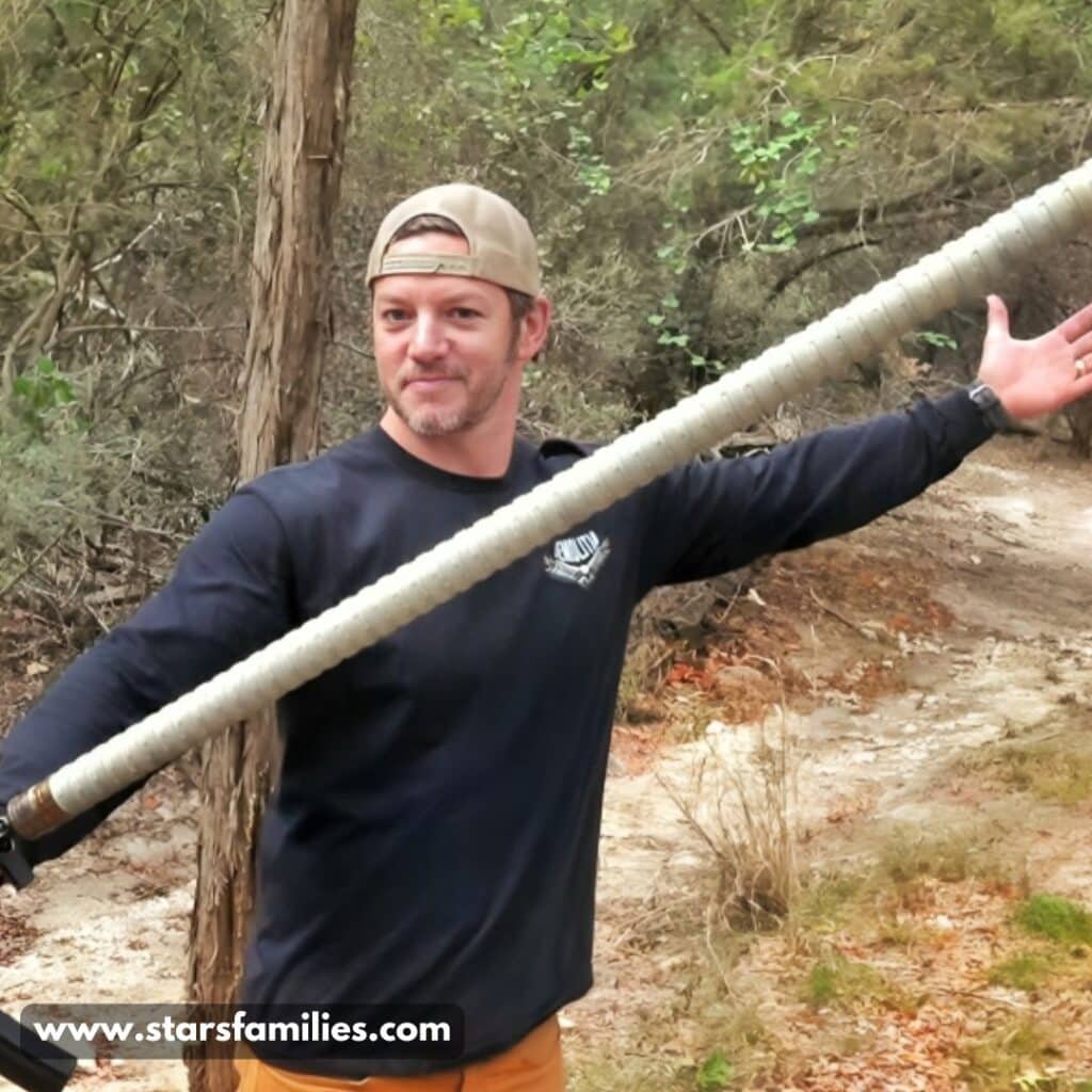 In a wooded outdoor setting, Matt Carriker, dressed in a dark long-sleeved shirt, beige cap, and orange pants, holds a large, wrapped pole or staff with both hands, arms extended outward