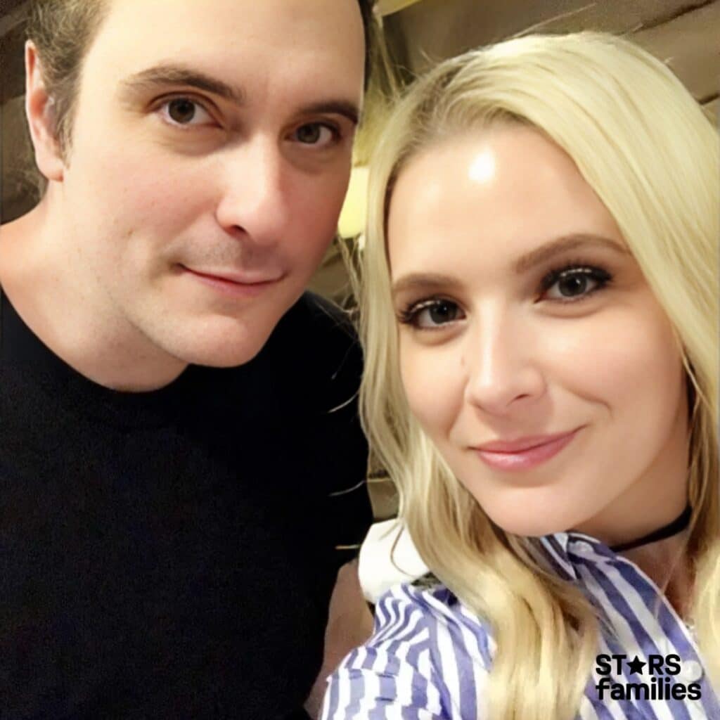 A close-up selfie of Benjamin Burnley and Rhiannon Napier, both smiling at the camera. Benjamin is wearing a black shirt and Rhiannon a blue and white striped top with a black choker.