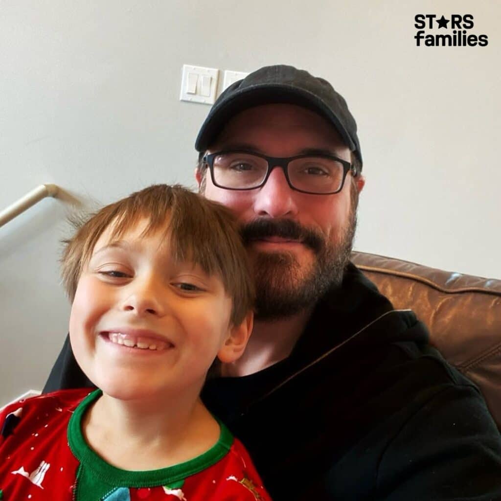 Benjamin Burnley taking a selfie with a young boy who is smiling broadly. Both wear casual attire and Benjamin has a beard and glasses, in a cozy home setting.