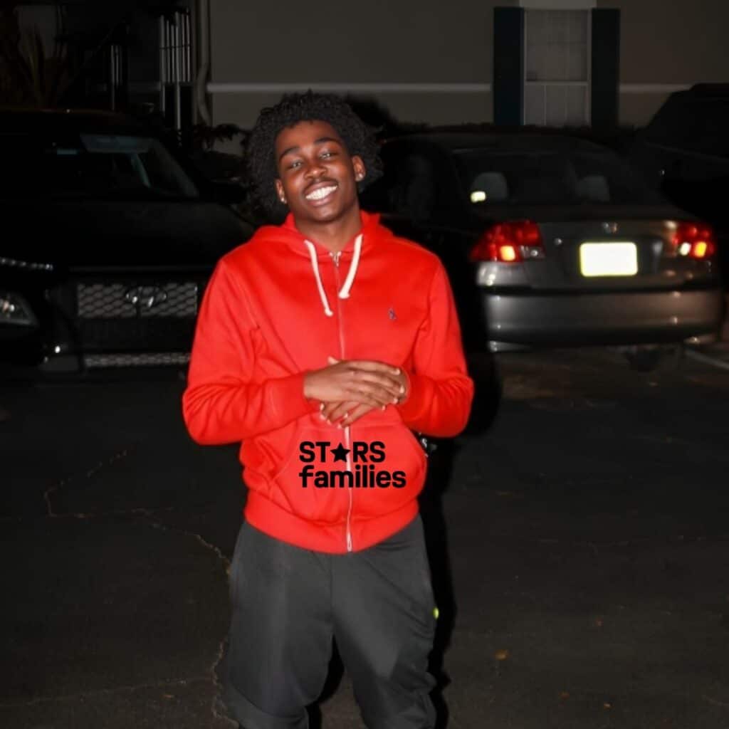 Kam Patterson, wearing a red hoodie and dark pants, stands outdoors at night. His face is blurred, and he stands in front of several parked cars and a building with a window.