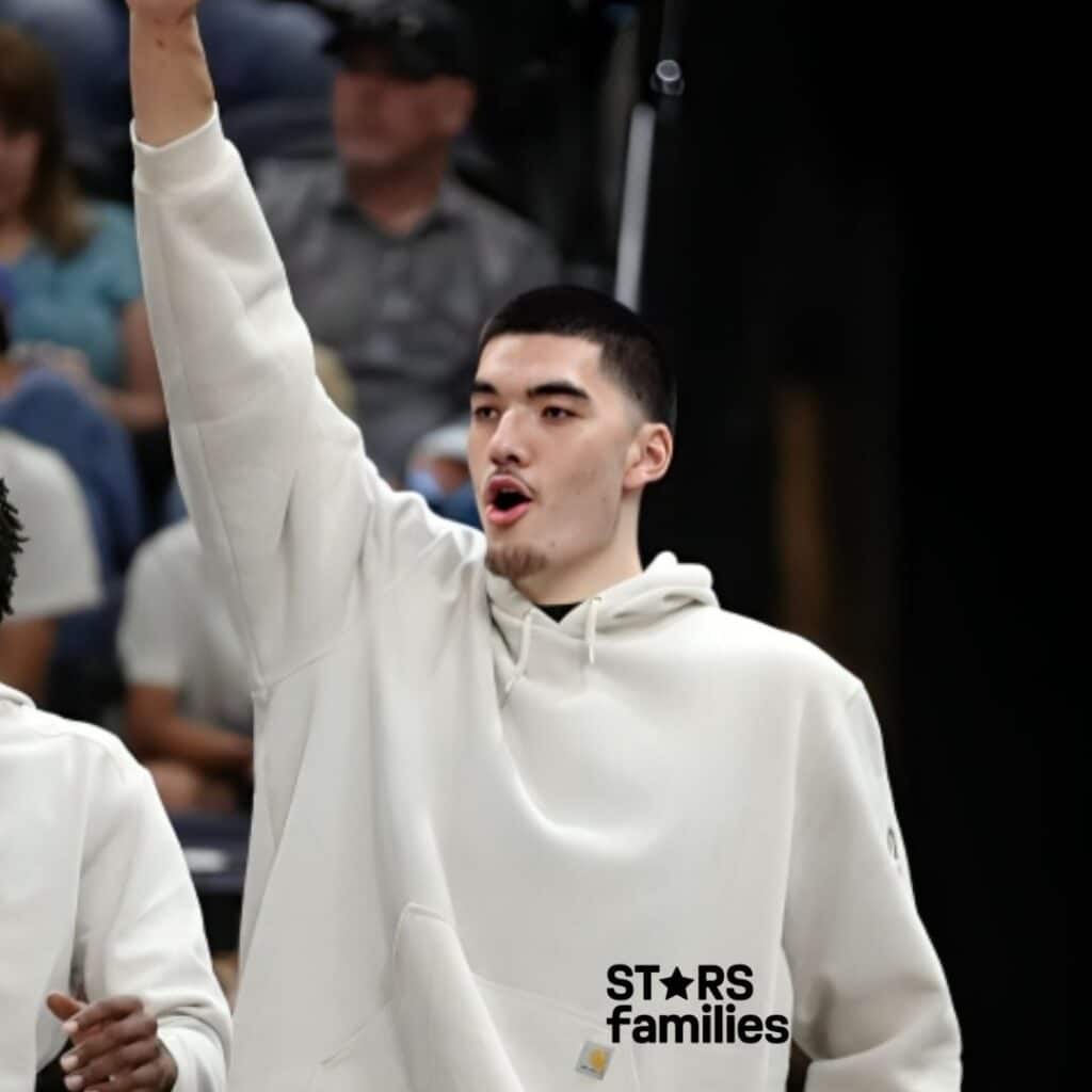 : Zach Edey, wearing a white hoodie, raises his arm at a public event. The background includes several people, possibly indicating a sports event or gathering.