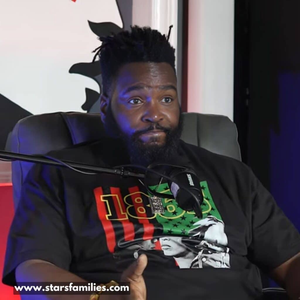 Dr. Umar Johnson sits in a chair with a microphone in front of him, suggesting he might be participating in a podcast or interview. He is wearing a black t-shirt with a design that includes the numbers "1865" in red and yellow, and an image of a person with a green background and red and white stripes