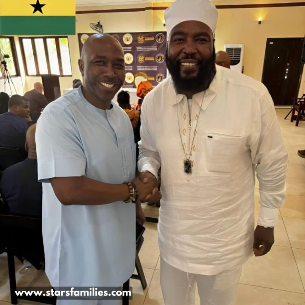 In an indoor formal event, Dr. Umar Johnson, wearing an all-white traditional outfit and cap, shakes hands with a person in a light blue traditional outfit.