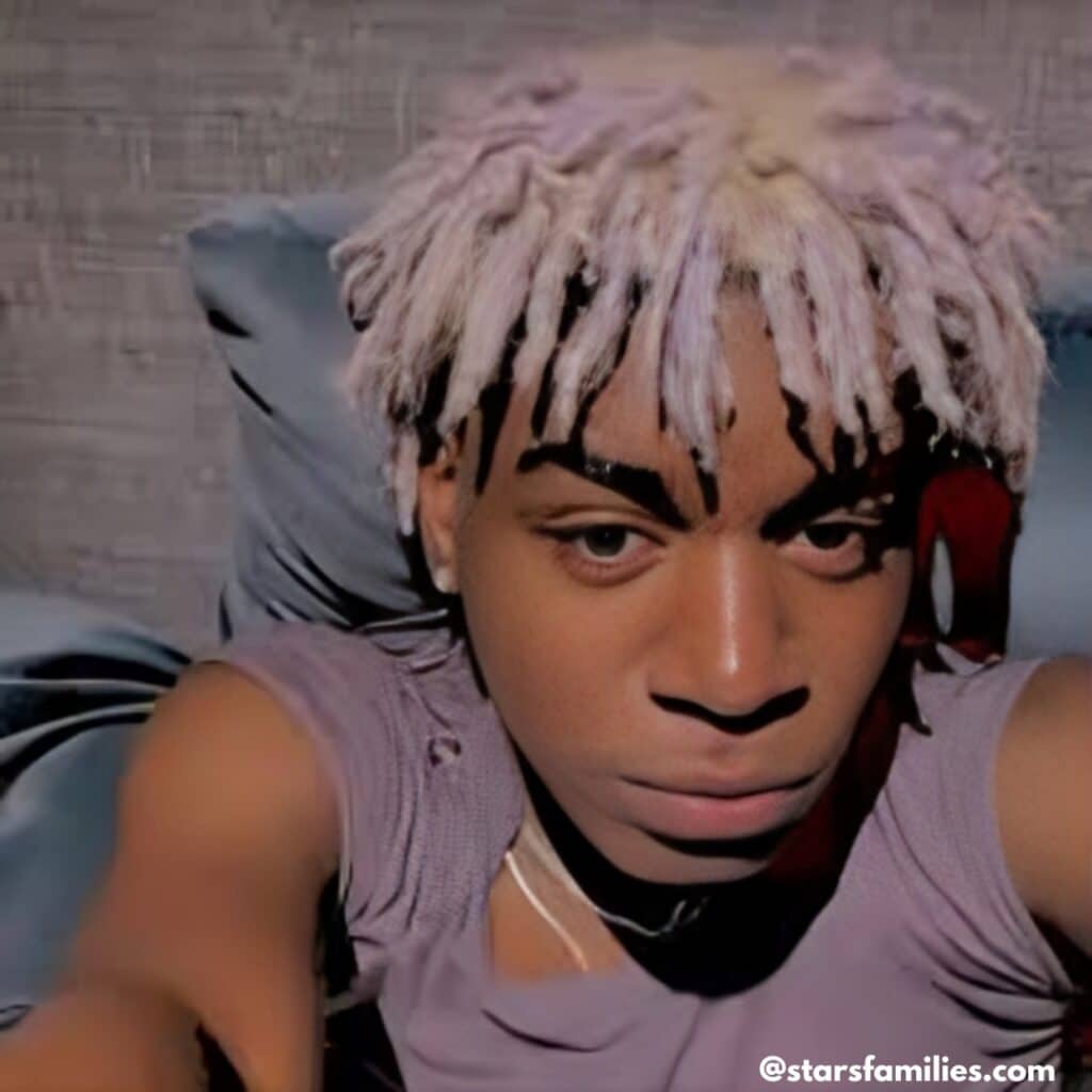 In a relaxed setting, Ken Carson, with bleached dreadlocks, is seated on a couch. He wears a sleeveless top, and the backdrop is a textured wall.