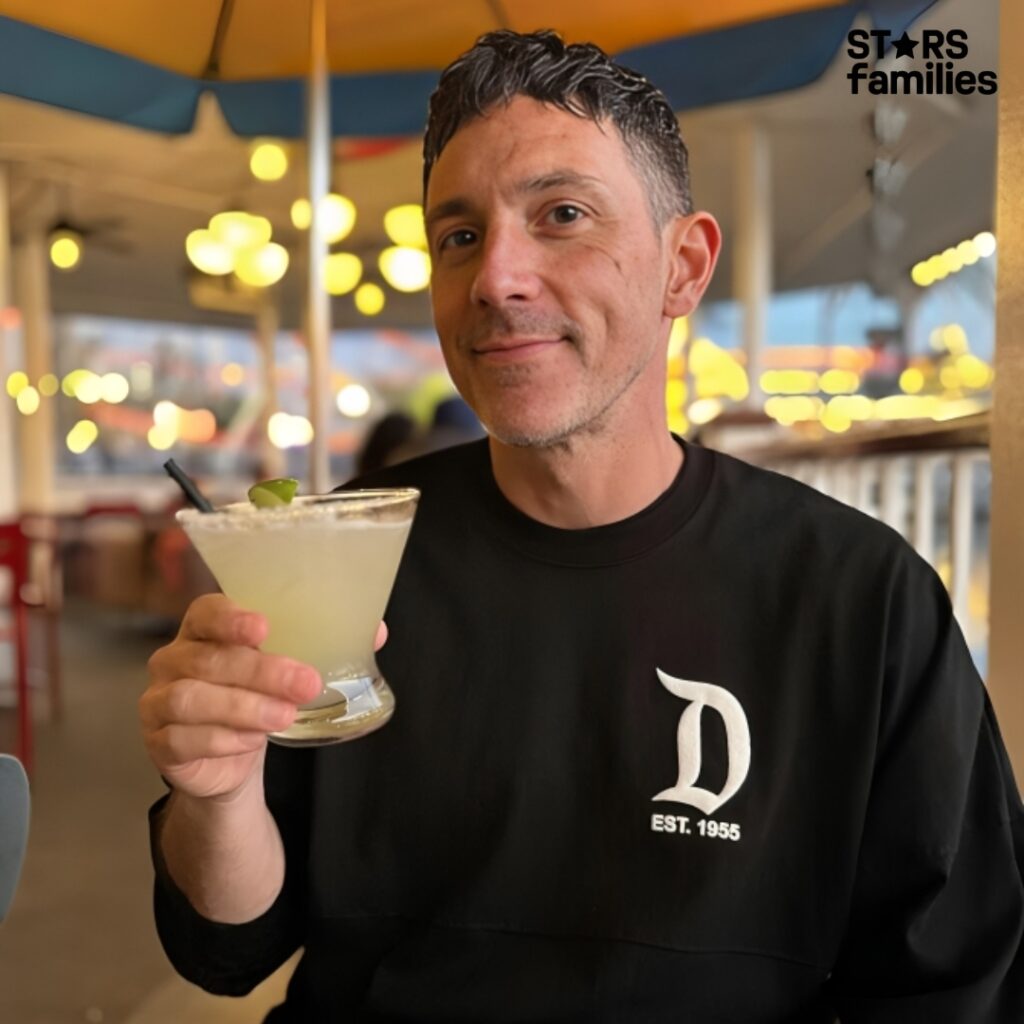 Steve Kazee in a black shirt, smiling while holding a drink, exuding a relaxed and cheerful vibe.