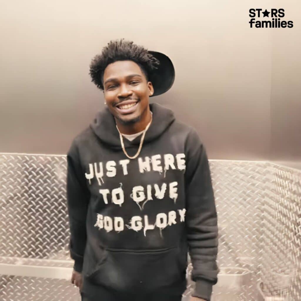 Kam Patterson stands in an elevator with metallic diamond plate walls. He is wearing a black hoodie with white text that reads, "JUST HERE TO GIVE GOD GLORY," and a gold chain necklace.