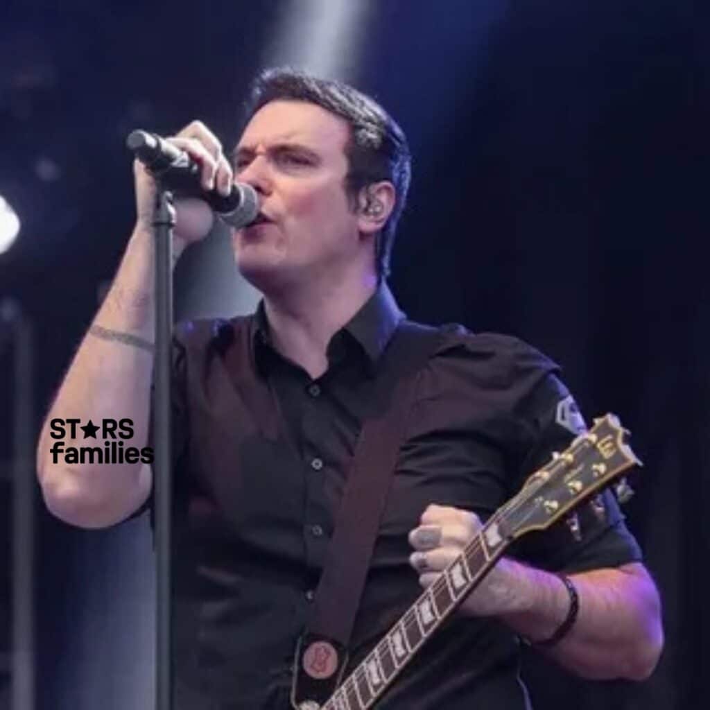 Benjamin Burnley performing on stage, singing into a microphone with a guitar strapped over his shoulder. He's wearing a dark, collared shirt and his left arm tattoos are visible.