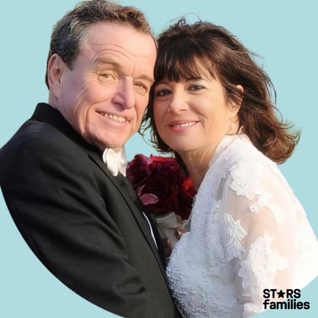 Jerry Mathers, dressed in a black suit with a white shirt and bow tie, stands beside his wife, who is wearing a white lace wedding dress and holding a bouquet of red flowers. They are outdoors, with palm trees in the background, suggesting a warm climate. The couple is captured in a joyful and significant moment, likely on their wedding day.