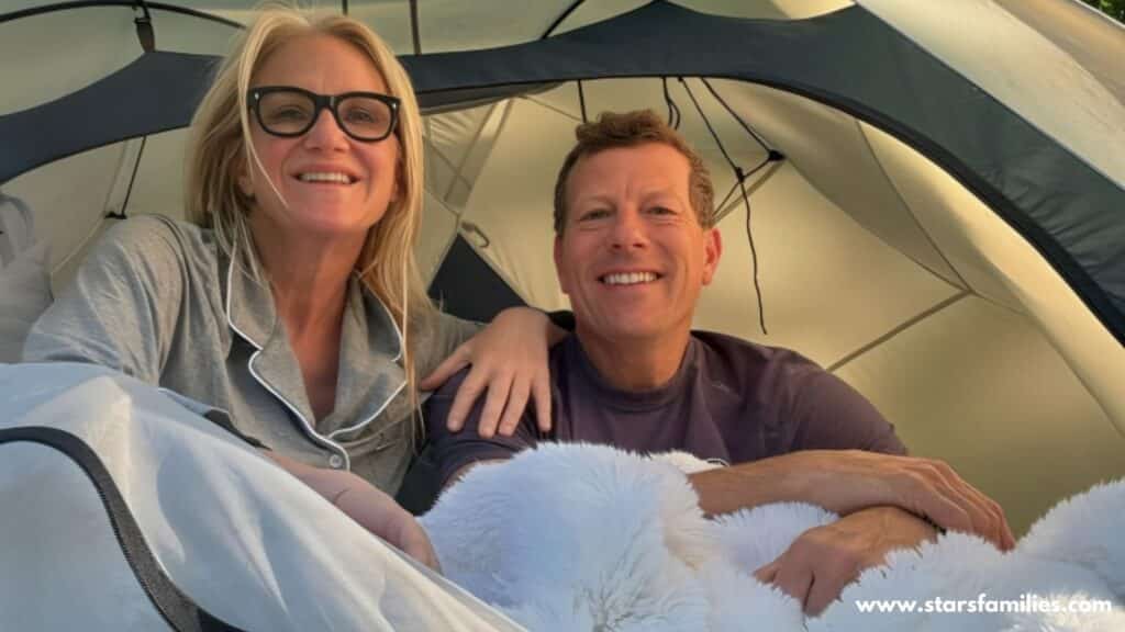 Mel Robbins, wearing grey pajamas with white piping, sits inside a tent. her husband, dressed in a dark-colored shirt, sits beside her. Both individuals have their faces obscured. The tent is set up outdoors, and there is a fluffy white blanket in front of them.