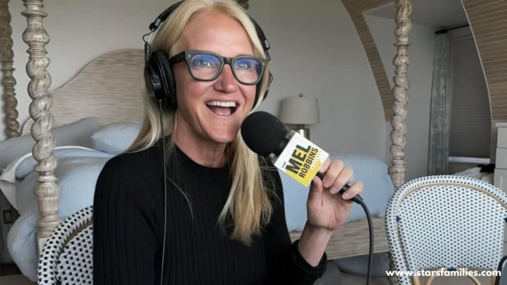 : Mel Robbins, seated in a room with a bed that has a decorative headboard and posts, holds a microphone with the label "The Mel Robbins Podcast" while wearing headphones. She is sitting comfortably and appears to be recording a podcast. The background includes a chair with a patterned cushion and a lamp on a nightstand.