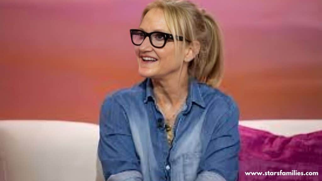 : Mel Robbins, sitting on a white couch with a pink pillow in the background, is wearing a blue denim shirt with the sleeves rolled up. She has a microphone clipped to her shirt. The background has a gradient of pink and orange colors.