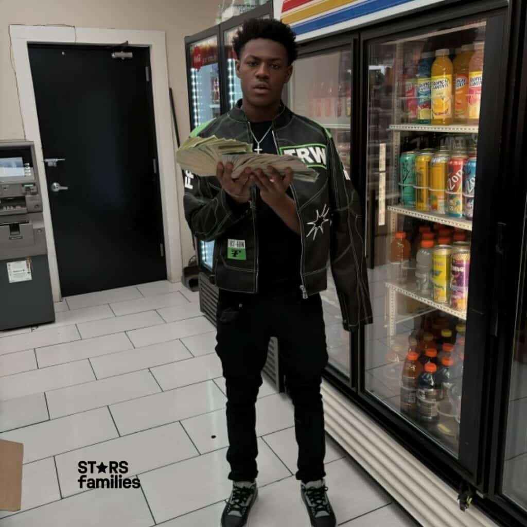 Baby Kia, wearing a black jacket with white text and graphics, black pants, and black and white sneakers, stands inside a store in front of refrigerated beverage displays. She is holding a large amount of cash in both hands. The background includes various drinks visible in the refrigerators, an ATM machine, and a black door.