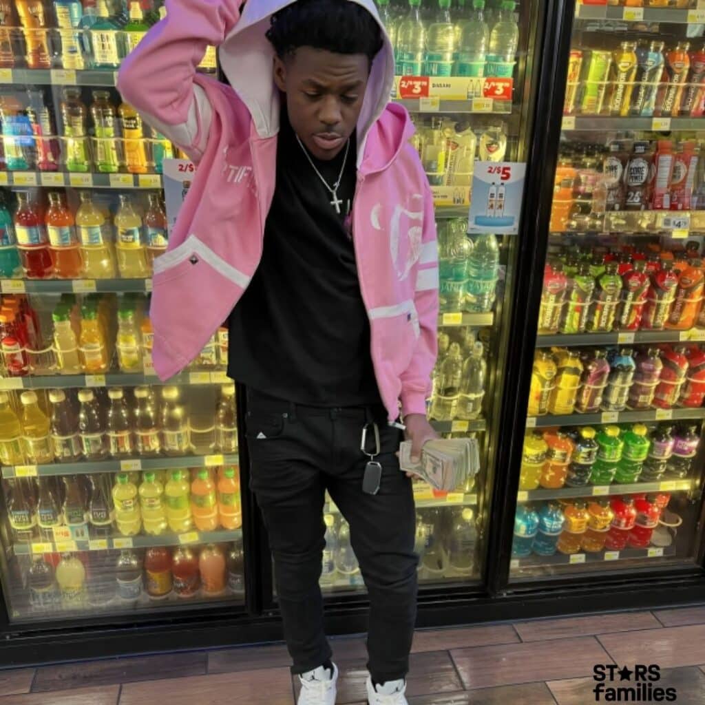 Baby Kia, wearing a pink hoodie with white stripes, a black shirt, black pants, and white sneakers, stands in front of a refrigerated section of a store stocked with various beverages. She holds a stack of cash in one hand and has a keychain attached to her pants. The background includes shelves filled with different colored bottles of drinks, with signs indicating prices such as "2/$3" and "2/$5."