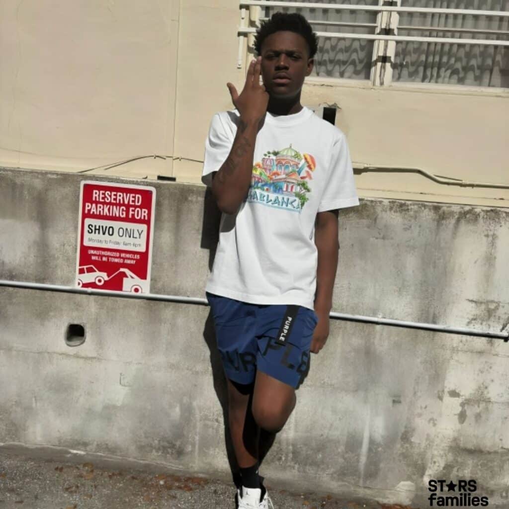 Baby Kia, wearing a white t-shirt with a colorful graphic that reads "CASABLANCA," blue shorts with the word "PURPLE" on the side, and white sneakers, stands against a beige concrete wall with a sign that says "Reserved Parking For SHVO Only." The background includes a window, andvisible wiring.