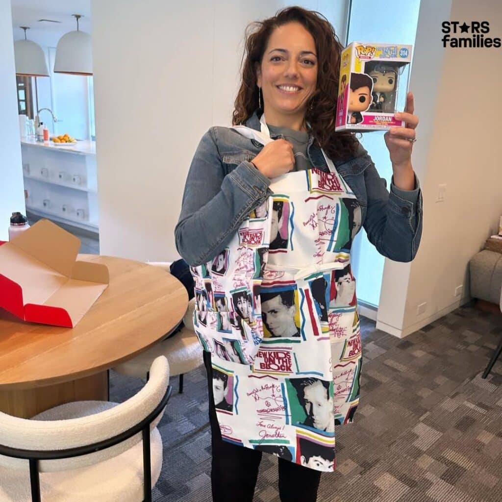 Sona Movsesian, standing in a modern kitchen or dining area, holds a Funko Pop! figure of Jordan Knight from New Kids on the Block. She is wearing an apron that features various images and text related to New Kids on the Block. The room includes a round wooden table with an open red and white box, white chairs, and a kitchen counter in the background.