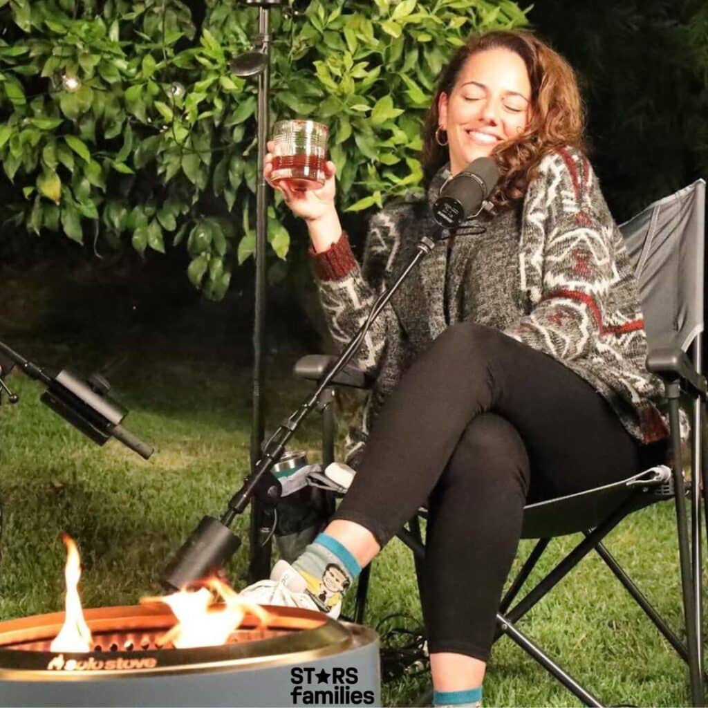 Sona Movsesian, sitting in a folding chair outdoors, holds a glass with a beverage in one hand. She is wearing a patterned sweater, black pants, and colorful socks with a character design. In front of her is a fire pit with visible flames, and the background features lush green foliage. She is also speaking into a microphone, suggesting a possible podcast or recording session.