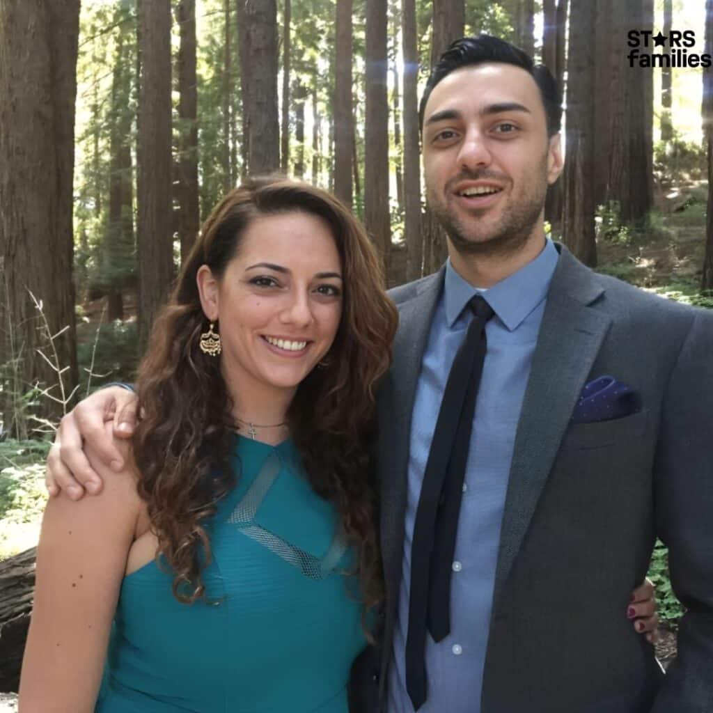 Sona Movsesian, wearing a teal dress with a cross necklace, stands outdoors in a forested area with tall trees in the background. She is accompanied by her husband, Tak Boroyan, who is dressed in casual attire. The setting appears to be a park or nature reserve during the daytime.