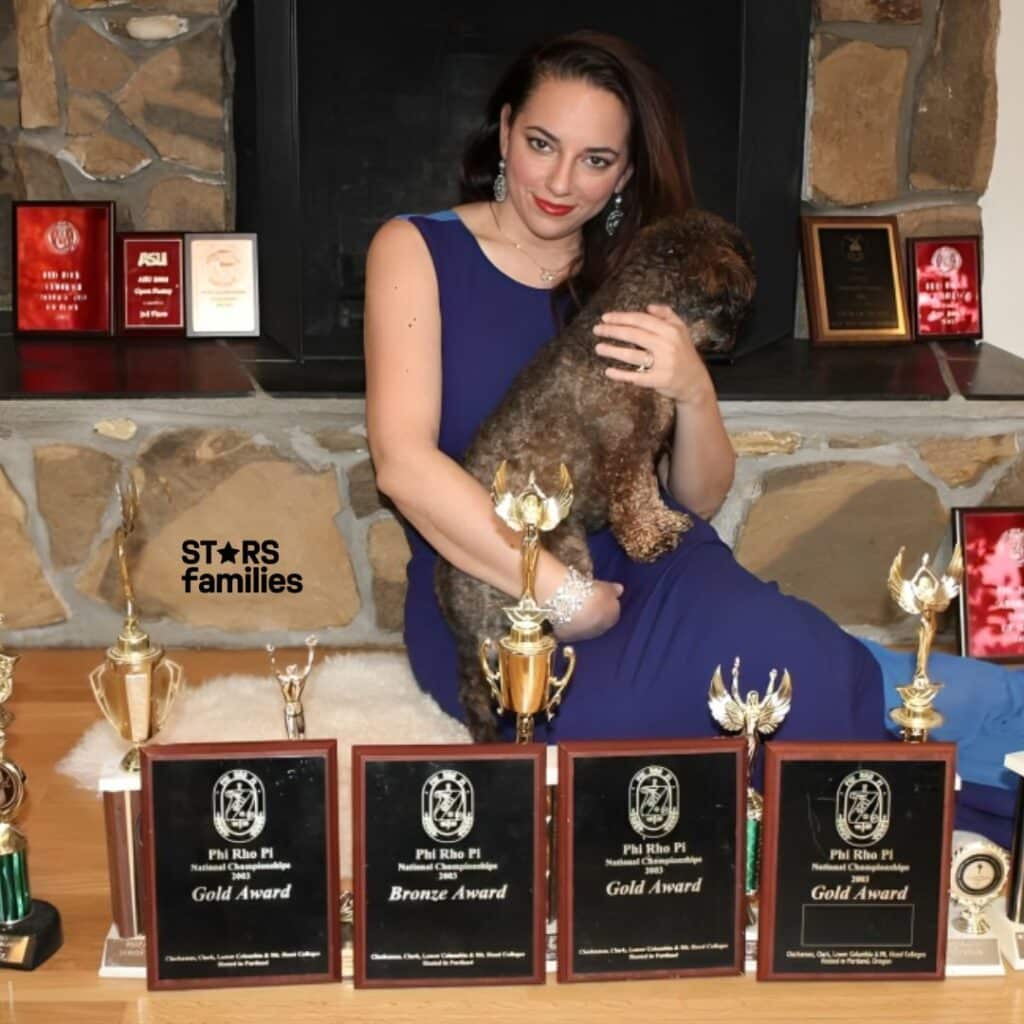 Sona Movsesian, wearing a blue dress, sits on the floor holding a small dog. In front of her are several trophies and plaques, including three Gold Awards, one Bronze Award, and various trophies of different sizes. The plaques are labeled "Phi Rho Pi National Championship" and display the years 2013 and 2015. The background features a stone wall with additional plaques and awards on a shelf.