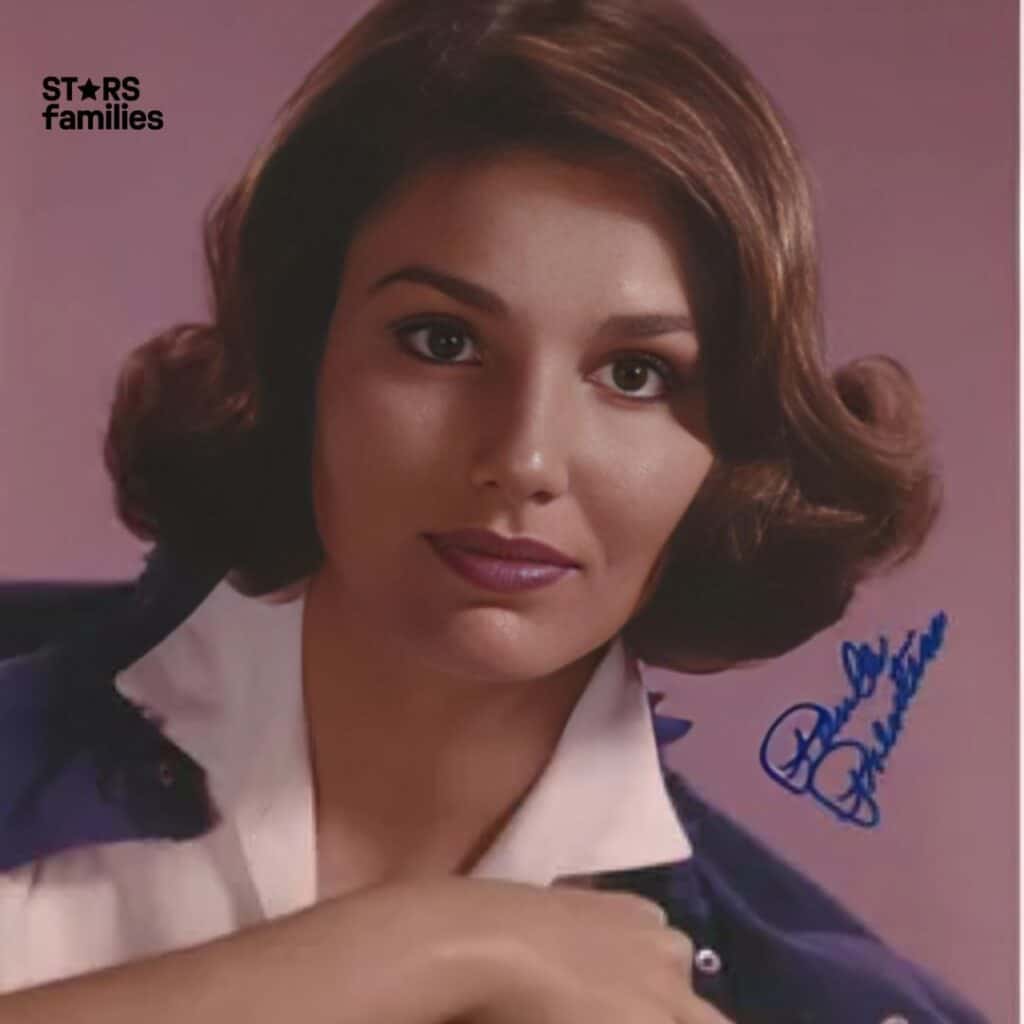 Paula Prentiss, with brown wavy hair, wearing a white collared shirt and a dark jacket, stands against a solid color background, possibly pink or purple.